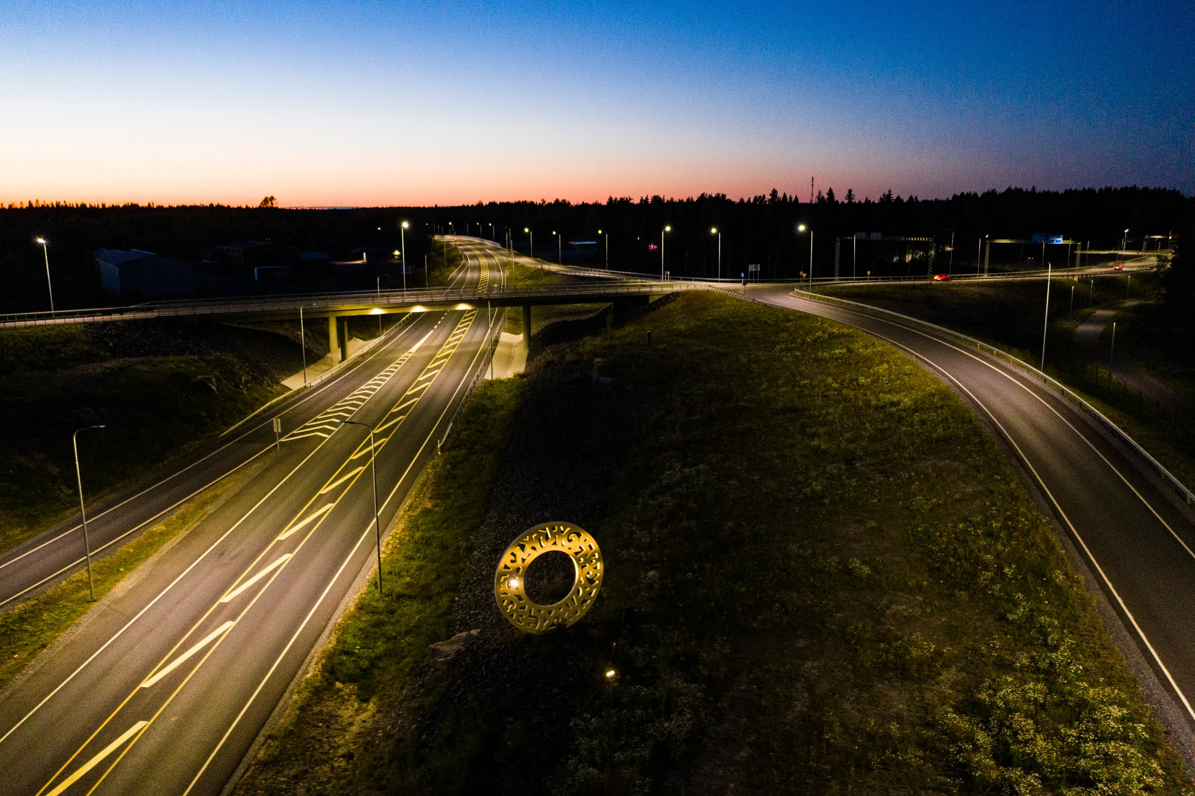 The Söörmarkku interchange from above at dusk: the bridge, the ramps and their lit poles, a lit sculpture in the green between the roads.